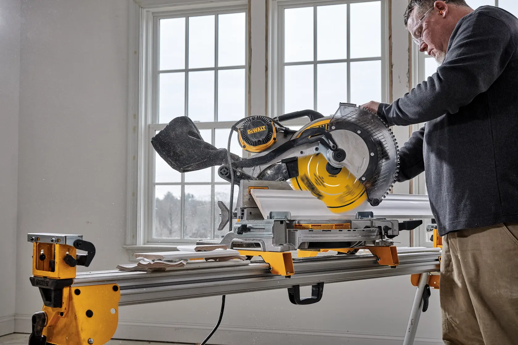 Uma pessoa usa uma serra de esquadria DEWALT para cortar uma peça de madeira dentro de uma oficina. O rosto da pessoa está desfocado e ela veste uma camisa xadrez e um boné. A serra é amarela e prateada, com uma mangueira de aspiração acoplada.