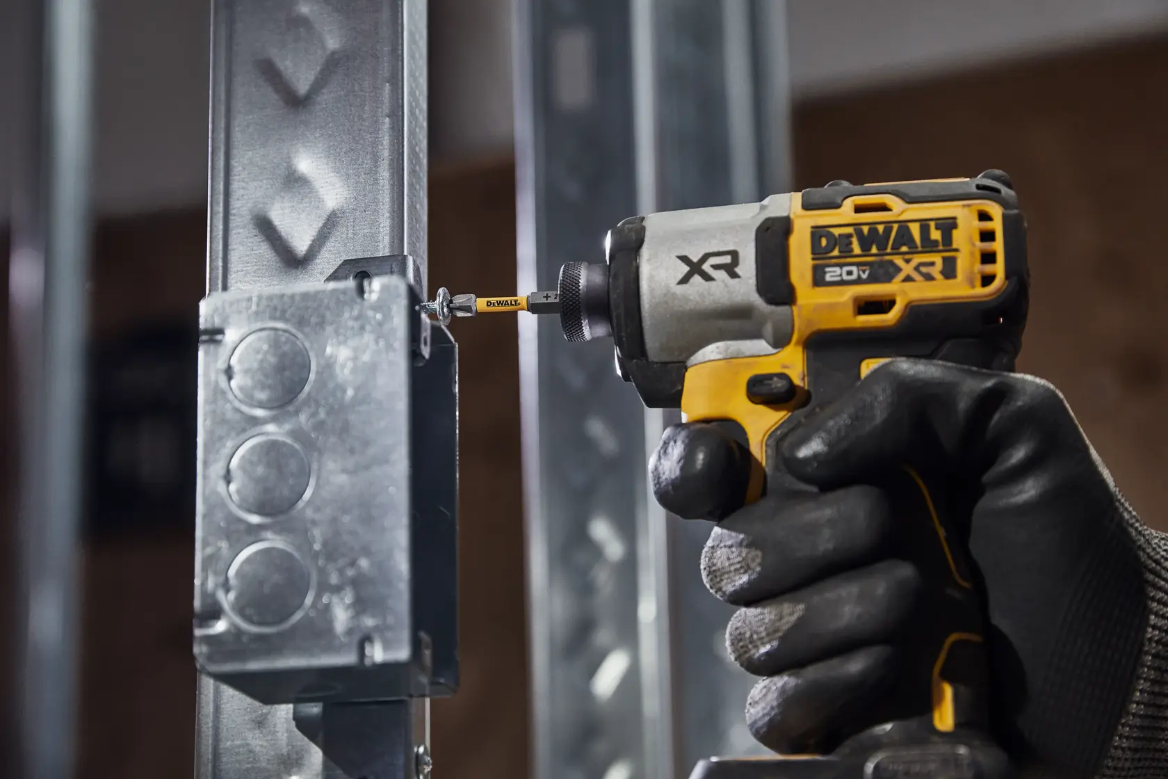 Uma mão com luva opera uma parafusadeira de impacto sem fio DEWALT 20V XR, apertando um parafuso em uma caixa elétrica de metal fixada em uma estrutura de aço.