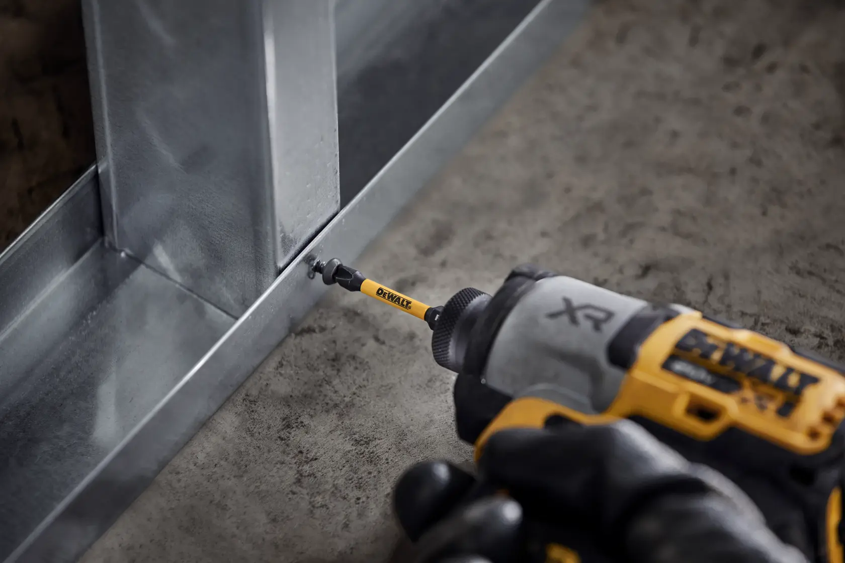 Close-up de uma furadeira sem fio DEWALT com uma ponteira para parafusar, instalando um parafuso em uma estrutura metálica sobre um piso de concreto.