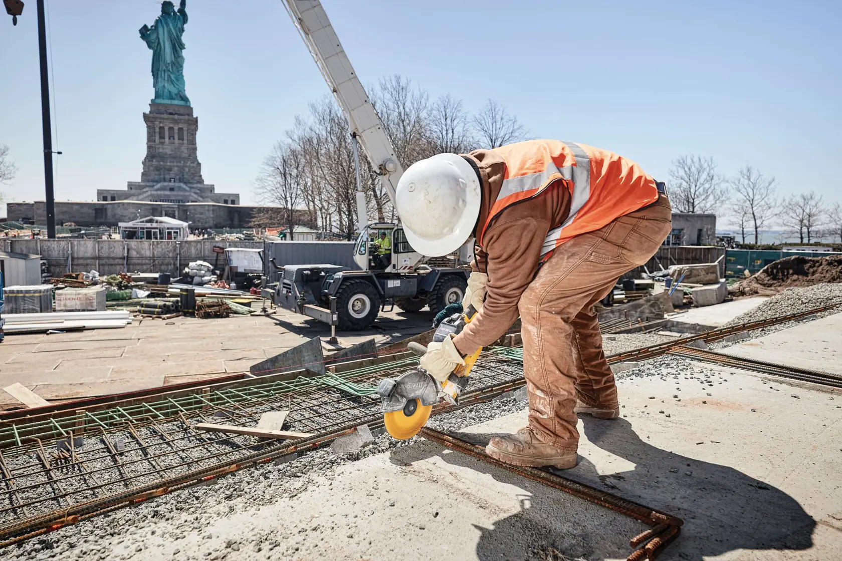 Um trabalhador da construção civil usando capacete de segurança, luvas e colete refletivo laranja utiliza uma ferramenta elétrica amarela para cortar vergalhão de metal em um canteiro de obras. A Estátua da Liberdade está visível ao fundo.