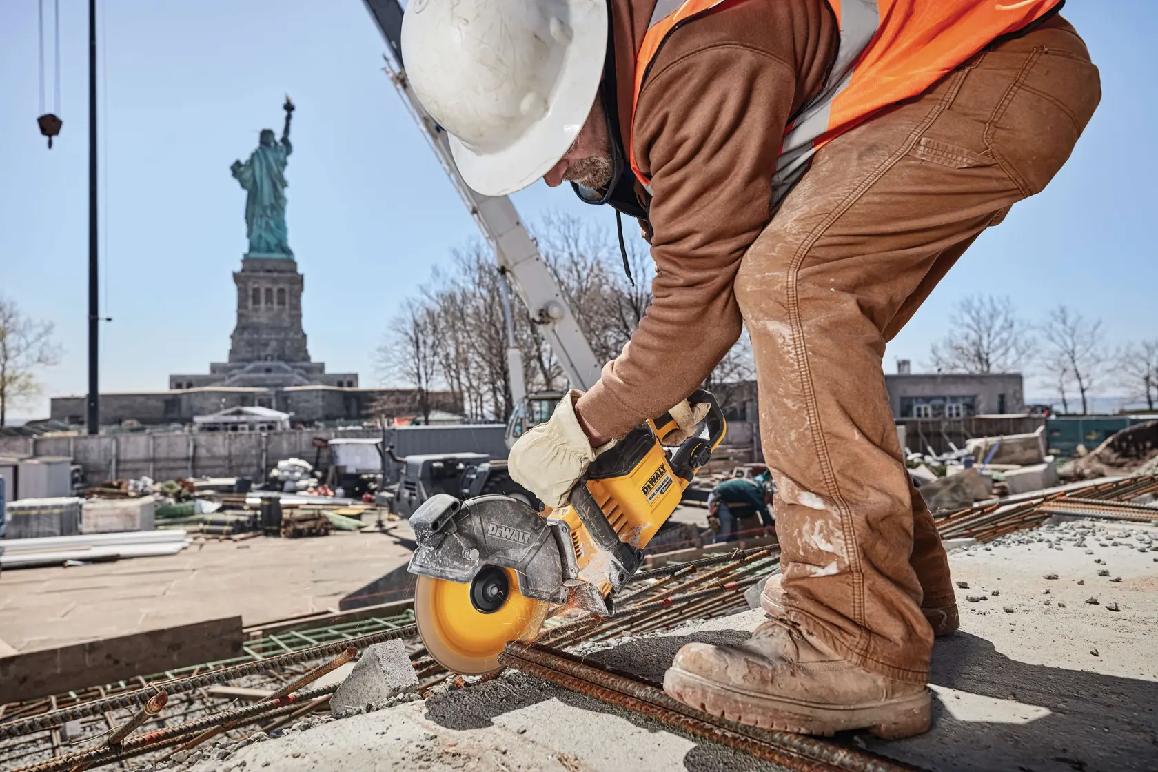 Uma pessoa usando capacete de construção e equipamentos de segurança utiliza uma ferramenta elétrica amarela DEWALT para cortar barras de metal em um canteiro de obras, com a Estátua da Liberdade visível ao fundo.