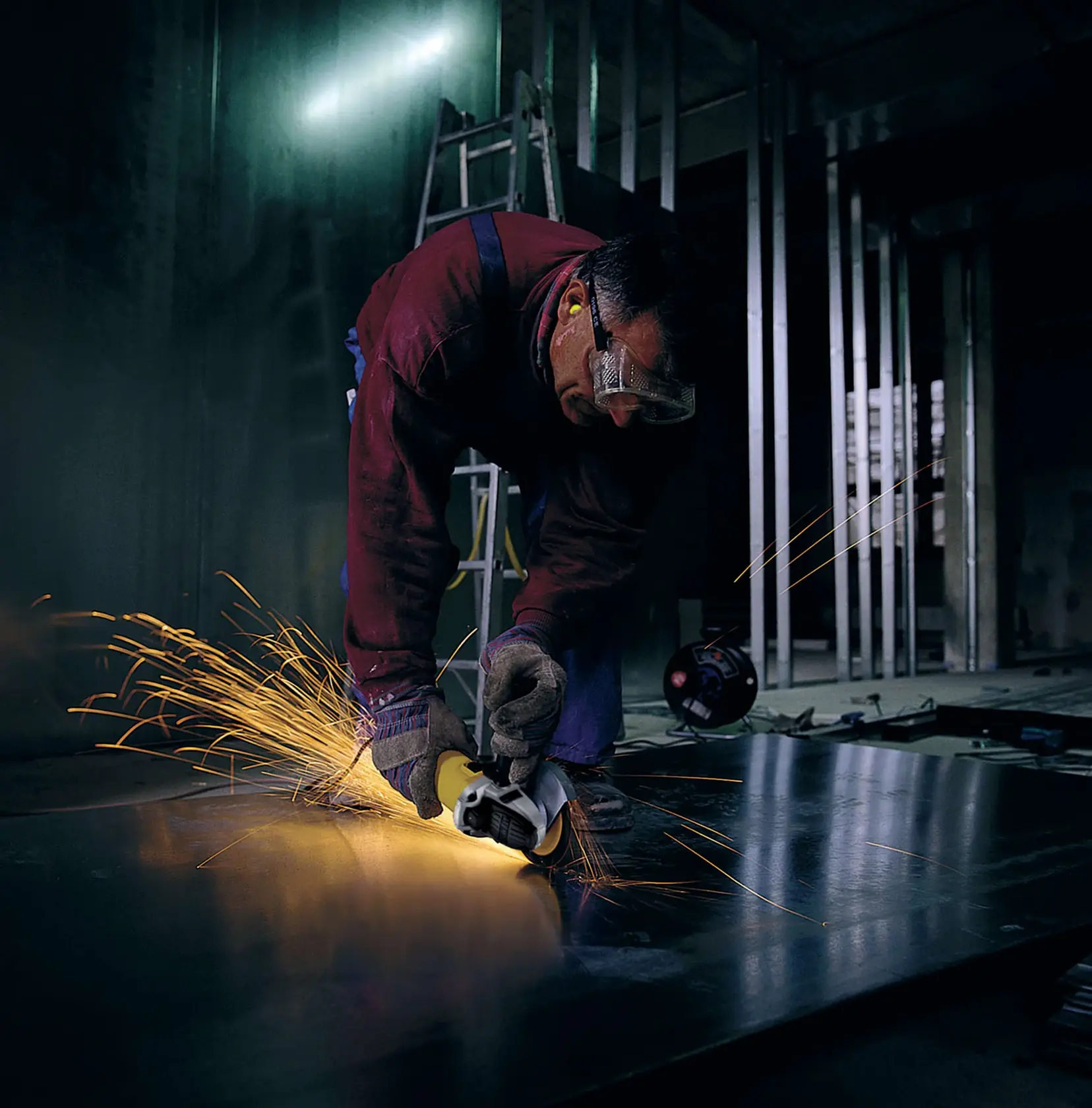 Uma pessoa usando luvas de proteção e protetores auriculares está utilizando uma lixadora angular DEWALT para cortar uma chapa de metal, produzindo faíscas em um ambiente industrial. O rosto da pessoa está desfocado.