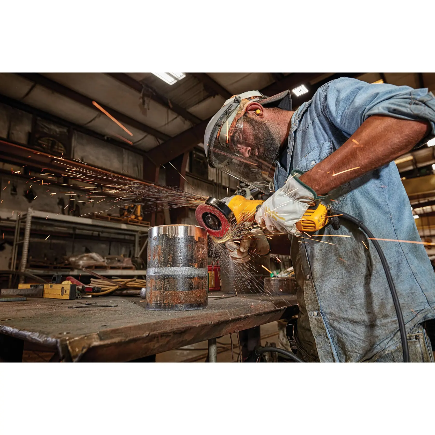 Um trabalhador usando equipamento de proteção, incluindo viseira, protetores auriculares, luvas e camisa jeans, utiliza uma esmerilhadora angular amarela DEWALT para lixar um cilindro de metal em uma bancada, com faíscas voando em uma oficina industrial.