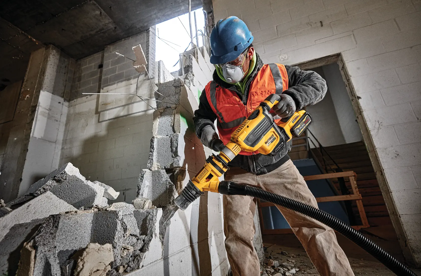 Um trabalhador da construção civil usando capacete azul e colete de segurança laranja utiliza uma ferramenta de demolição DEWALT com acessório de extração de pó para quebrar uma parede de concreto dentro de um edifício parcialmente demolido.
