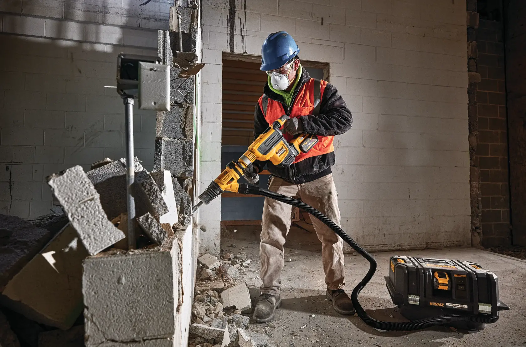Um trabalhador de construção com capacete azul e colete de segurança laranja usa uma ferramenta de demolição DEWALT para quebrar uma parede de concreto. Há entulho no chão e uma caixa de equipamentos DEWALT com uma mangueira conectada à ferramenta.
