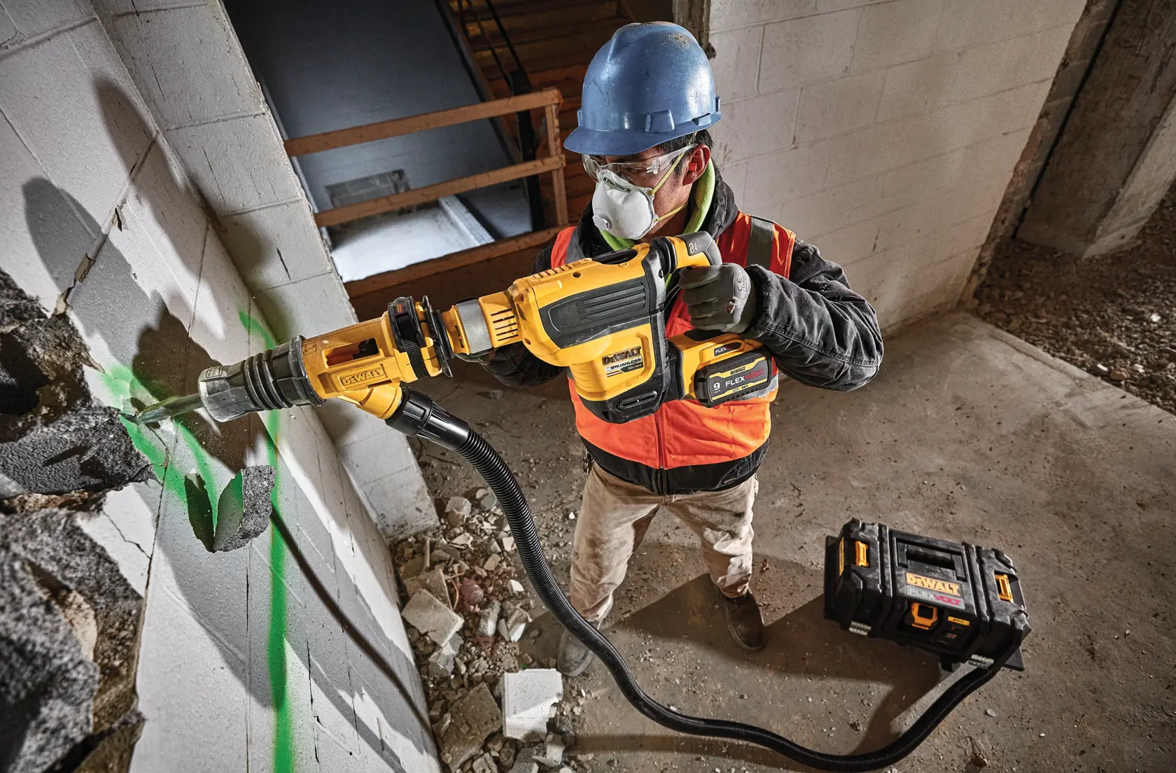 Um trabalhador da construção civil usando capacete azul e colete laranja está utilizando uma ferramenta elétrica DEWALT para perfurar uma parede de concreto. A ferramenta está conectada por uma mangueira a uma caixa de ferramentas DEWALT preta e amarela no chão. Marcas de tinta verde estão visíveis na parede.