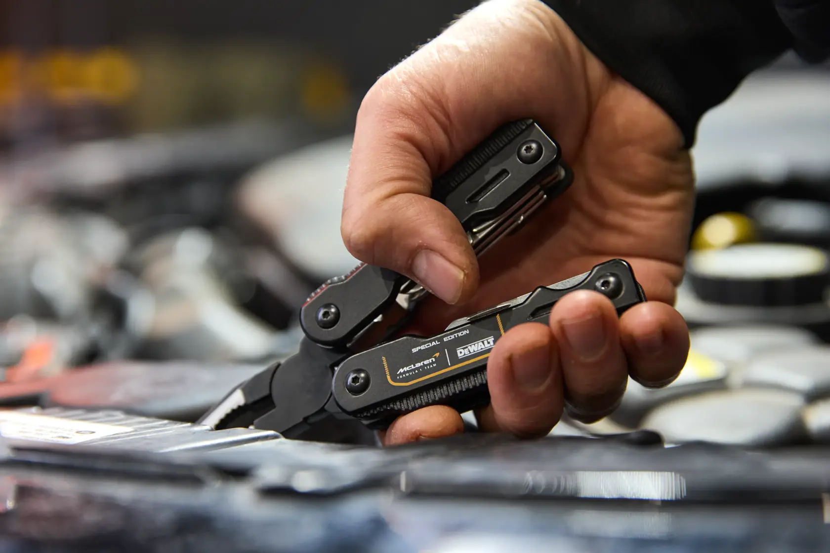 Mão usa alicate multifuncional preto DEWALT, com várias ferramentas visíveis, em oficina.