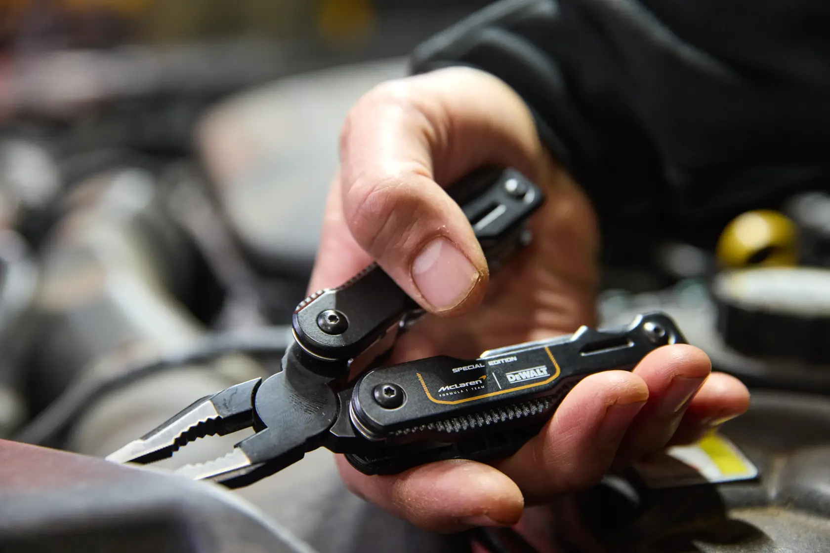 Mão segurando um multitool preto DEWALT com alicate, usado em trabalho mecânico numa oficina.