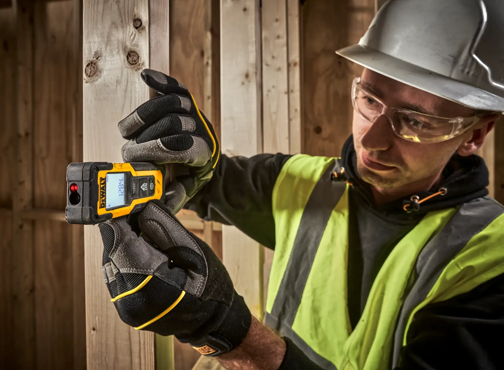 Uma mão com luva segurando um medidor de distância a laser DEWALT com uma tela digital exibindo os resultados das medições, em um ambiente de construção com vigas de madeira ao fundo.