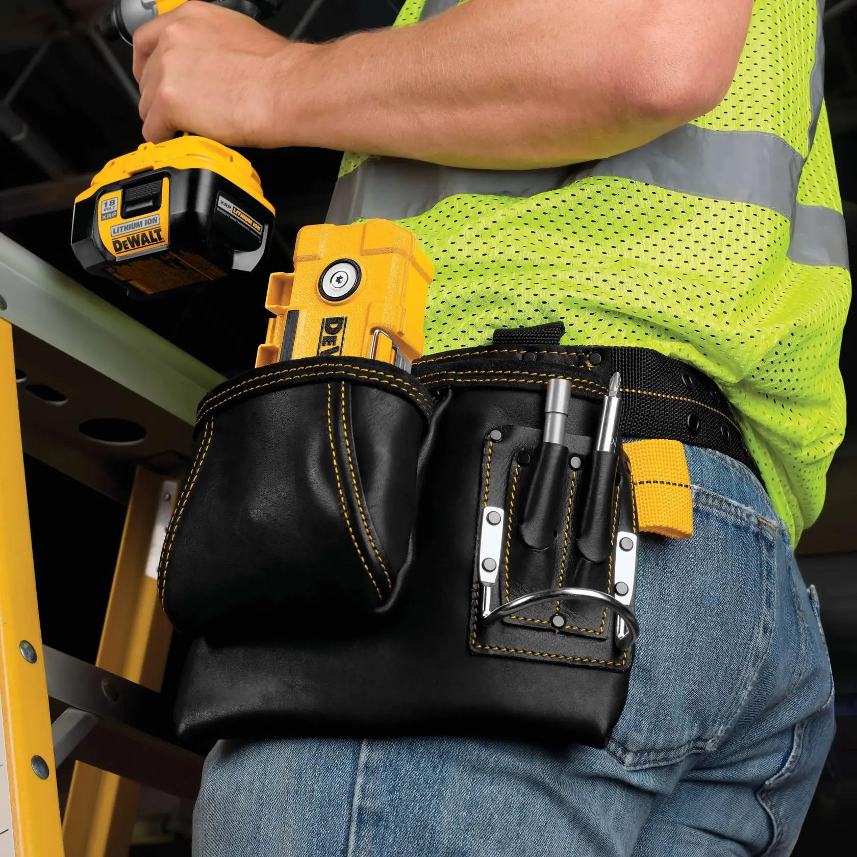 Vista de perto de uma pessoa usando um cinto de ferramentas de couro preto com costura amarela, contendo diversas ferramentas e acessórios DEWALT. A pessoa também está segurando uma ferramenta elétrica sem fio DEWALT e veste jeans azuis e um colete de segurança amarelo brilhante. Uma escada amarela está visível ao fundo.
