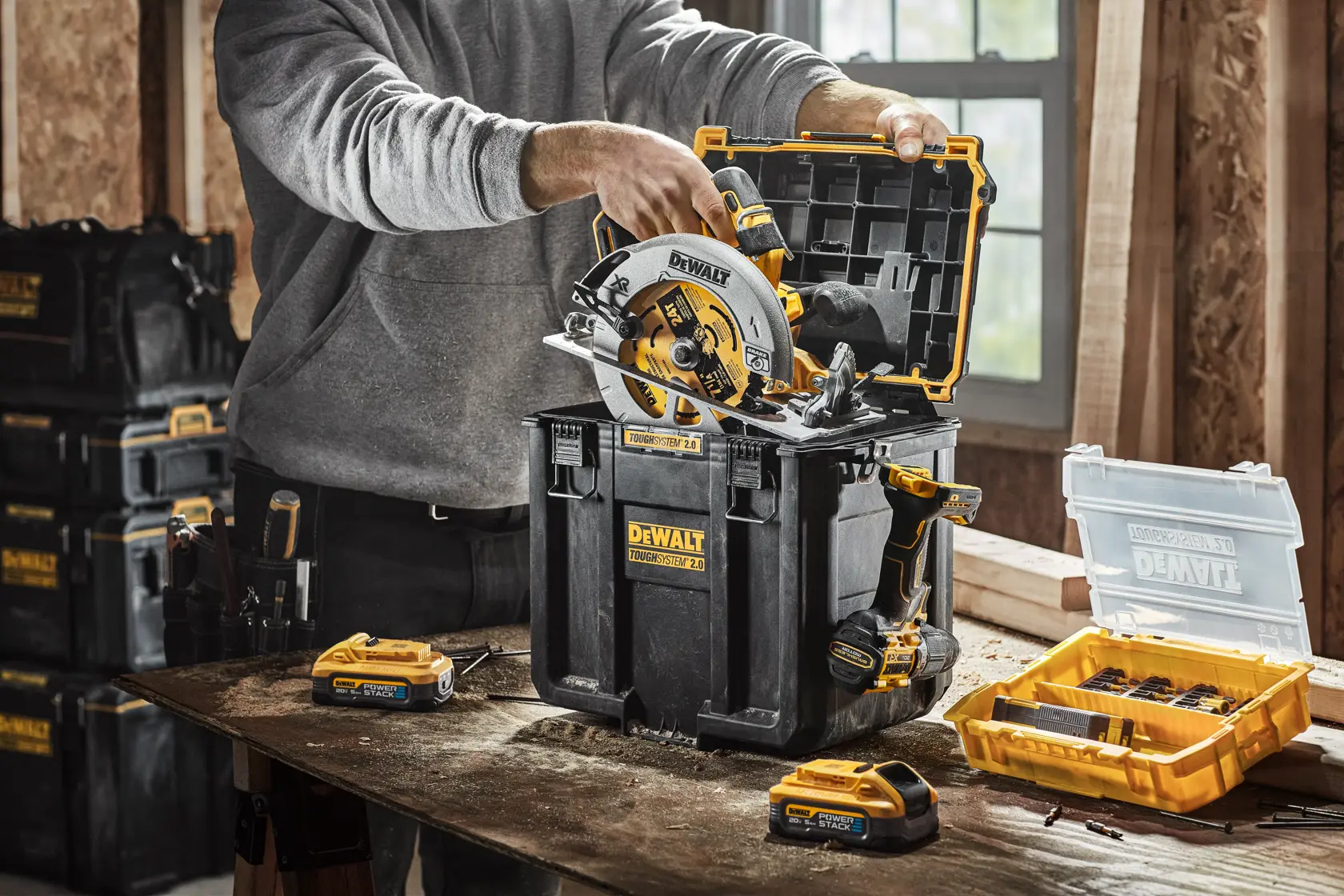 Uma pessoa organiza ferramentas em uma caixa de armazenamento empilhável DEWALT com organizador com tampa e 4 divisórias removíveis. A caixa é preta com detalhes amarelos e faz parte de um sistema de armazenamento modular com rodas.
