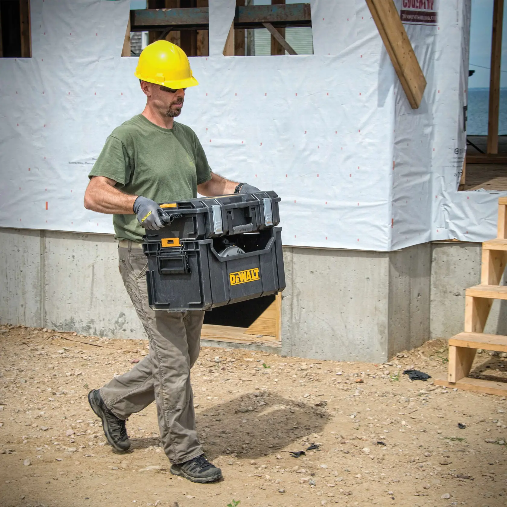 Uma pessoa usando capacete amarelo, luvas e camiseta verde está carregando uma caixa de ferramentas preta DEWALT (SKU: DWST08205) em um canteiro de obras com uma estrutura parcialmente construída ao fundo.
