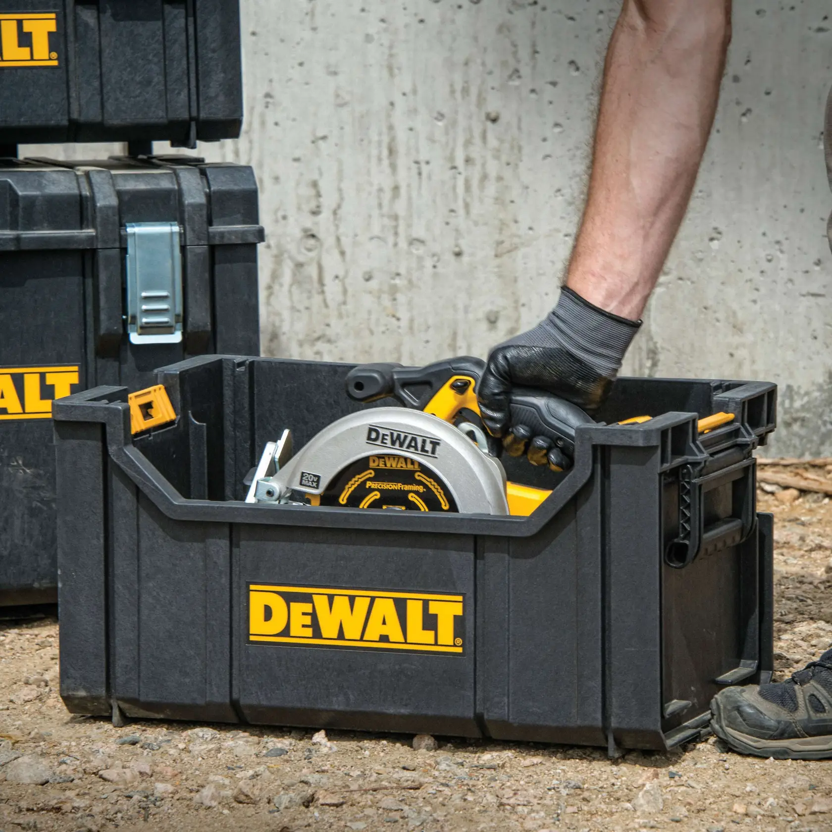Uma caixa de armazenamento preta da marca DEWALT é mostrada em um local de construção. Uma pessoa com luvas está colocando uma serra circular e outras ferramentas dentro da caixa. Mais caixas de armazenamento DEWALT aparecem ao fundo.