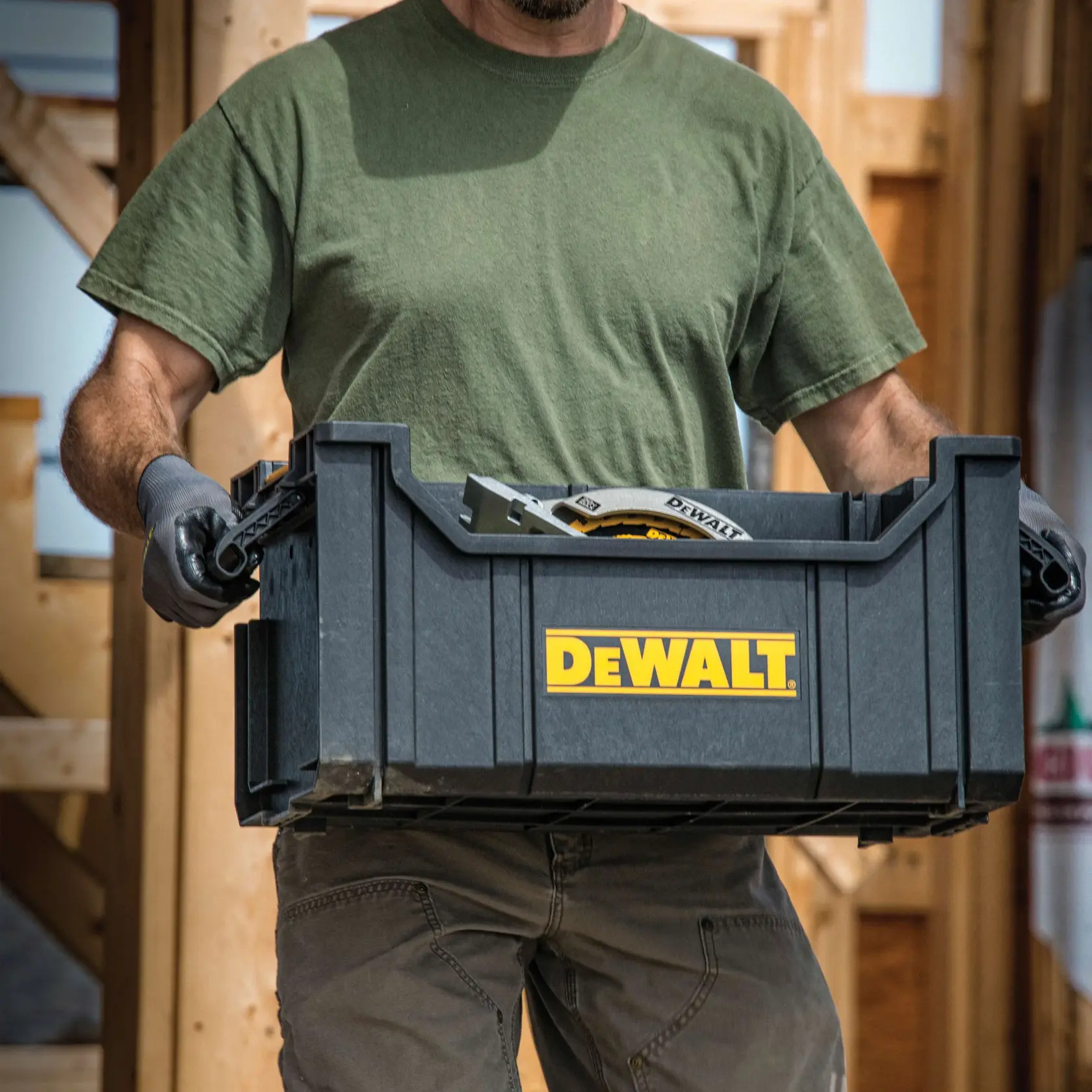Uma pessoa vestindo uma camiseta verde e luvas está carregando um recipiente de armazenamento de plástico preto DEWALT cheio de ferramentas. O logotipo DEWALT é claramente visível na frente do recipiente. A cena parece ser em um canteiro de obras com estrutura de madeira ao fundo.