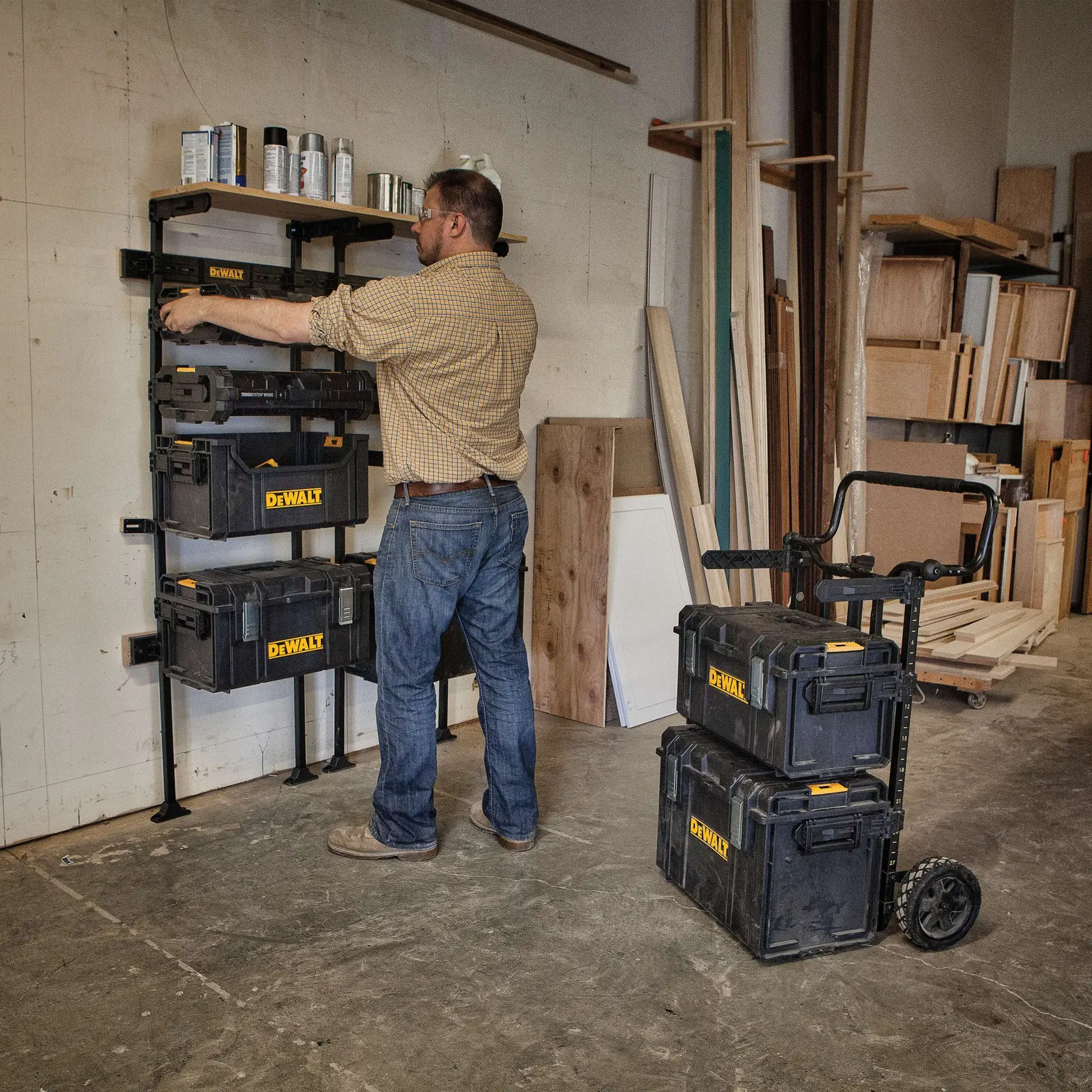 Uma pessoa está organizando caixas de armazenamento DEWALT em uma prateleira montada na parede de uma oficina. Outras caixas DEWALT estão empilhadas em um carrinho com rodas próximo. O ambiente possui várias placas de madeira e prateleiras ao fundo.