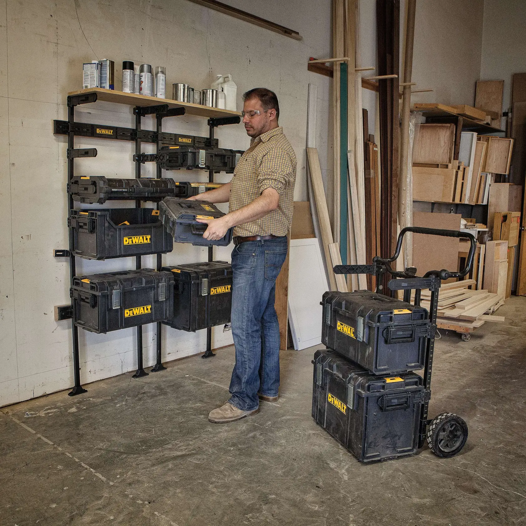 Um homem em uma oficina está organizando caixas de ferramentas empilháveis DEWALT, colocando uma em um suporte de armazenamento montado na parede. Várias caixas de ferramentas pretas e amarelas DEWALT estão armazenadas no suporte, e duas estão empilhadas em um carrinho com rodas próximo. O espaço de trabalho contém tábuas de madeira e prateleiras com diversos suprimentos.