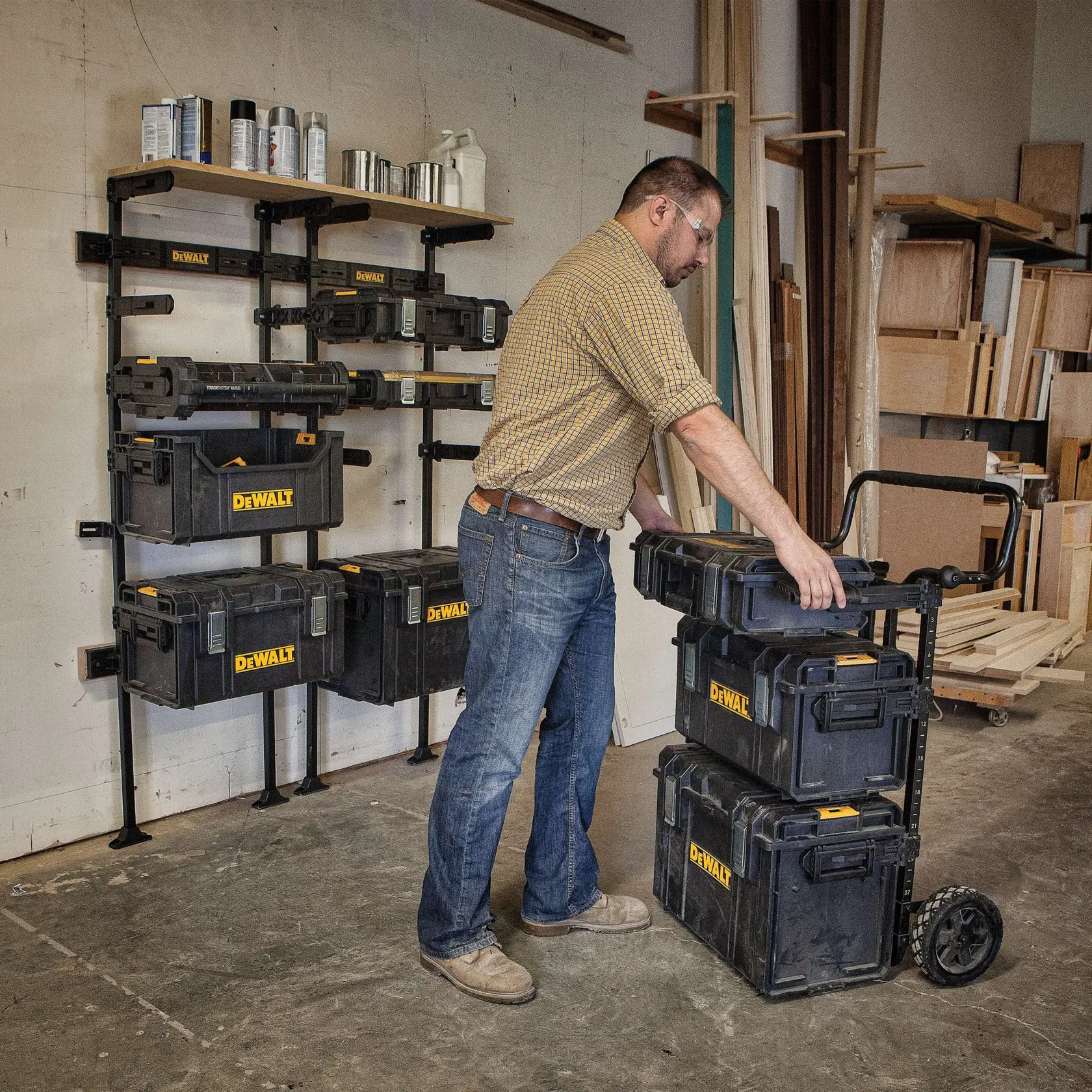 Uma pessoa em uma oficina organiza caixas de armazenamento DEWALT, algumas montadas em uma estante de parede e outras empilhadas em um carrinho com rodas. A oficina contém materiais de madeira e prateleiras ao fundo.