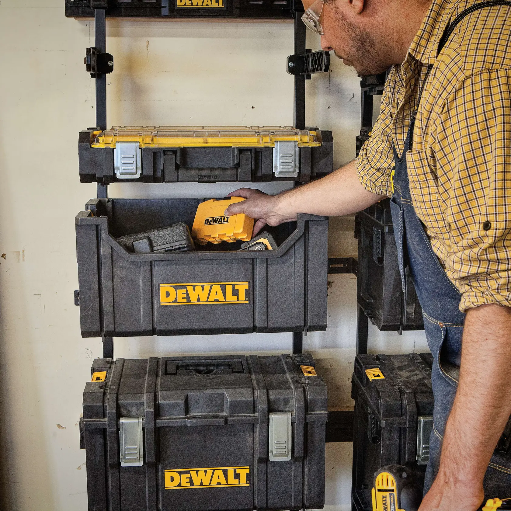 Uma pessoa com camisa xadrez e macacão organiza caixas de armazenamento DEWALT fixadas na parede. A pessoa está colocando uma ferramenta elétrica DEWALT dentro de uma das caixas. As caixas são pretas com logotipos amarelos da DEWALT.