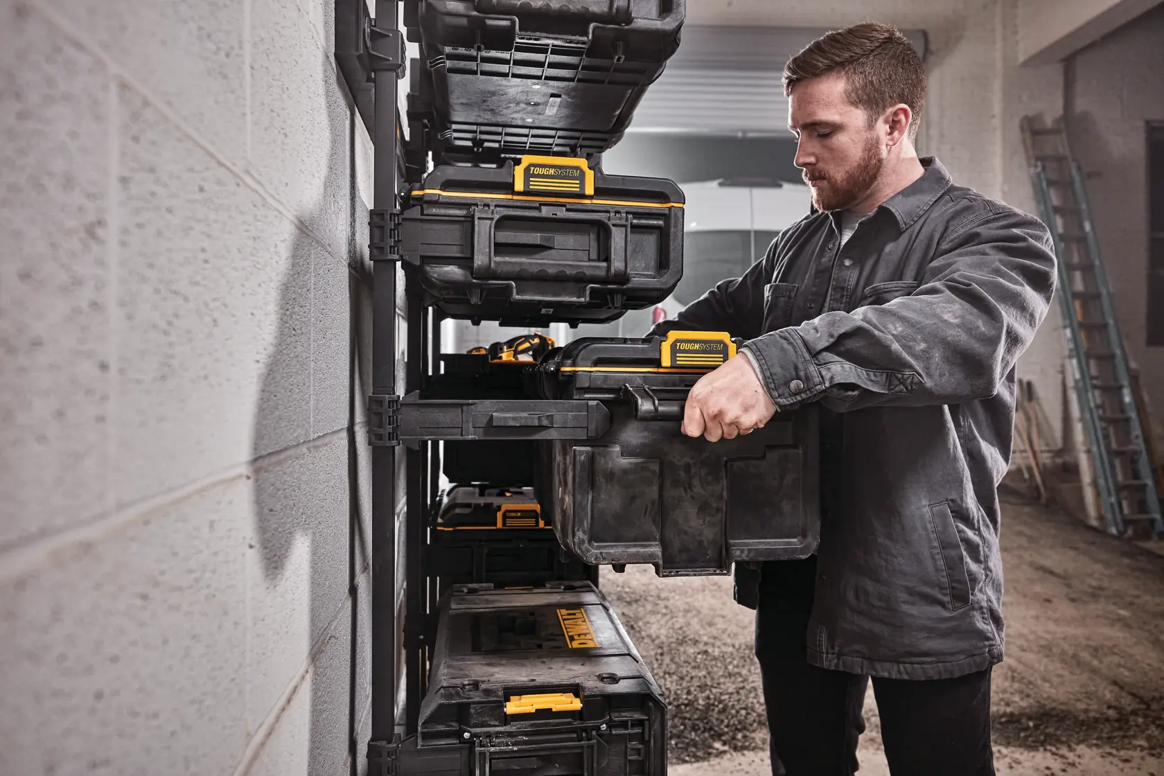 Uma pessoa está organizando caixas de armazenamento DEWALT em uma prateleira montada na parede, em uma oficina ou garagem. O rosto da pessoa está desfocado e ela veste uma jaqueta de trabalho escura.