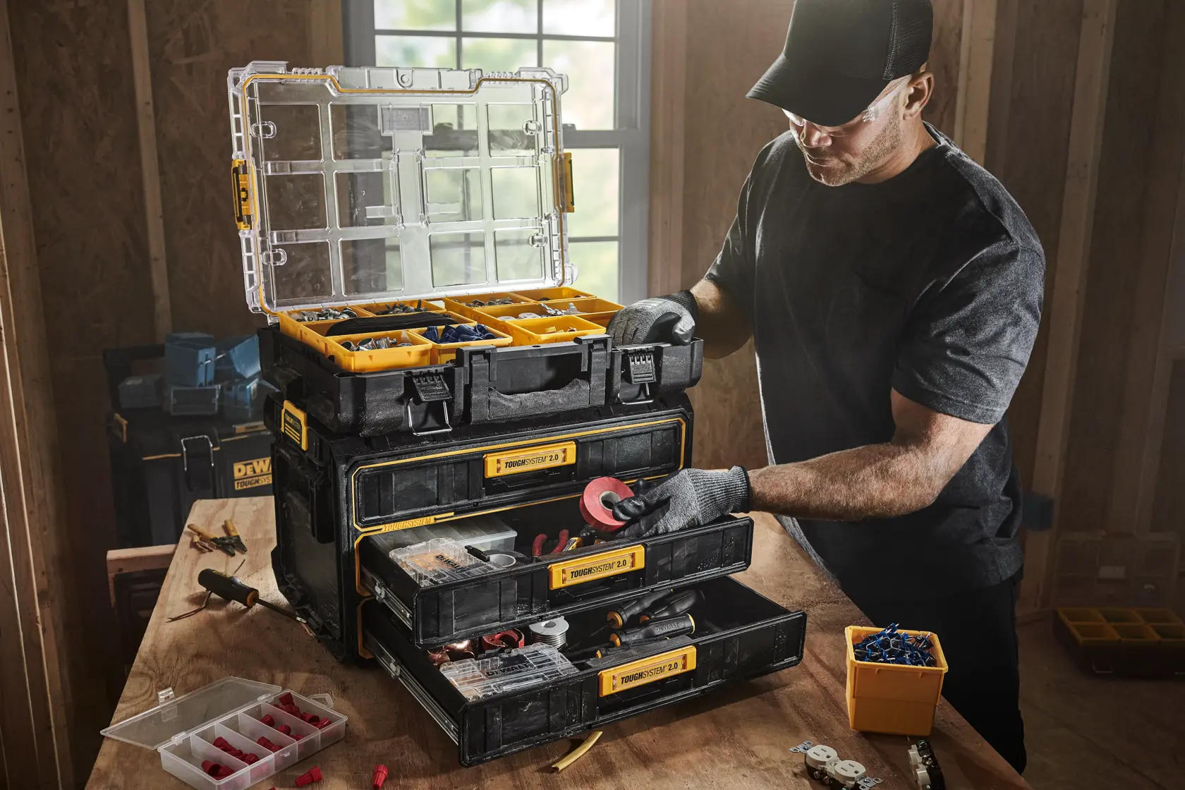 Uma pessoa utiliza uma unidade de armazenamento DEWALT ToughSystem 2.0 com três gavetas rasas e um compartimento superior aberto, organizando ferramentas e acessórios em uma bancada de trabalho em uma oficina.