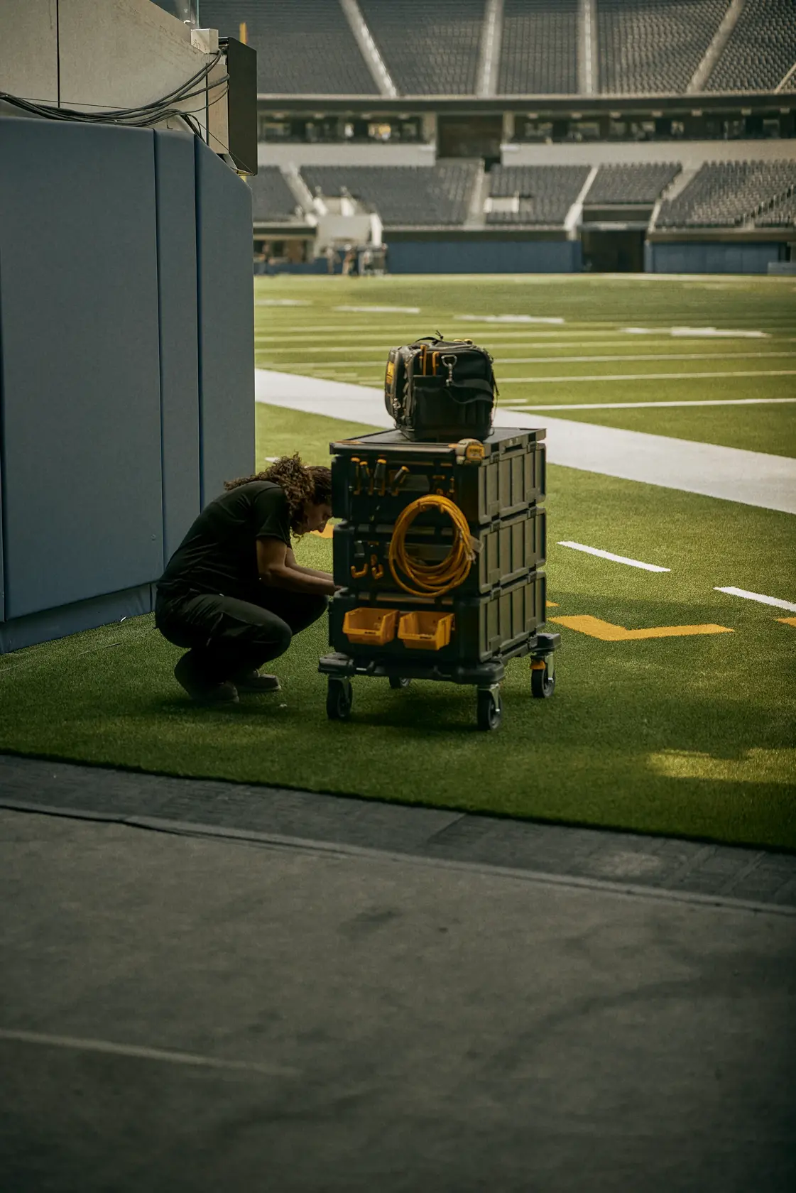 Uma pessoa agachada ao lado de um carrinho de armazenamento móvel DEWALT com caixas de ferramentas empilhadas e um cabo amarelo enrolado, localizado em um campo de estádio coberto.
