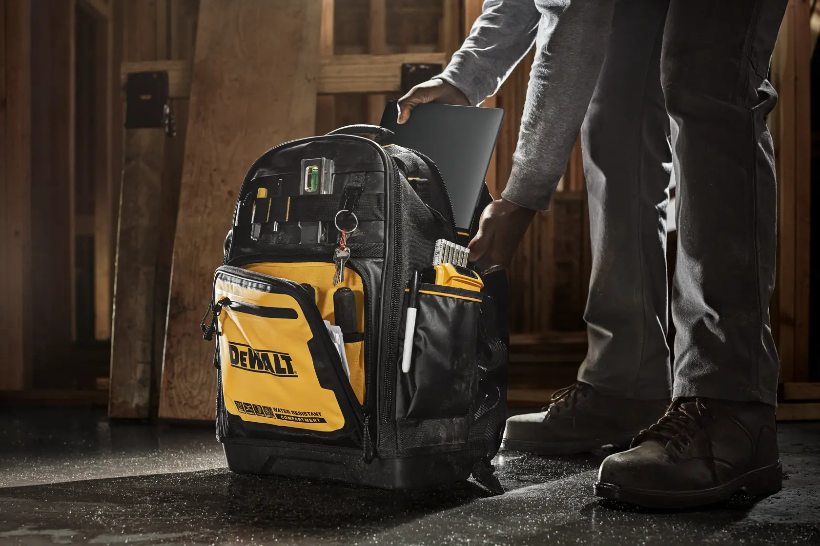 Uma mochila de ferramentas DEWALT com vários compartimentos e ferramentas organizadas, sendo usada em um canteiro de obras. Uma pessoa está colocando um tablet na mochila.