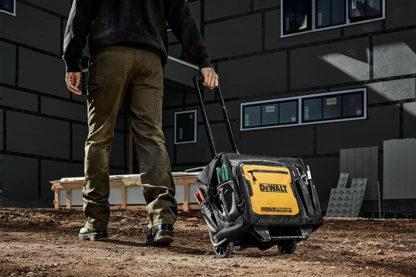 Uma pessoa puxa uma bolsa de ferramentas DEWALT com alça extensível em um canteiro de obras ao ar livre. A bolsa é preta e amarela, com vários bolsos externos cheios de ferramentas. O texto promocional abaixo destaca a otimização do armazenamento, 27 bolsos e um compartimento para objetos pessoais.
