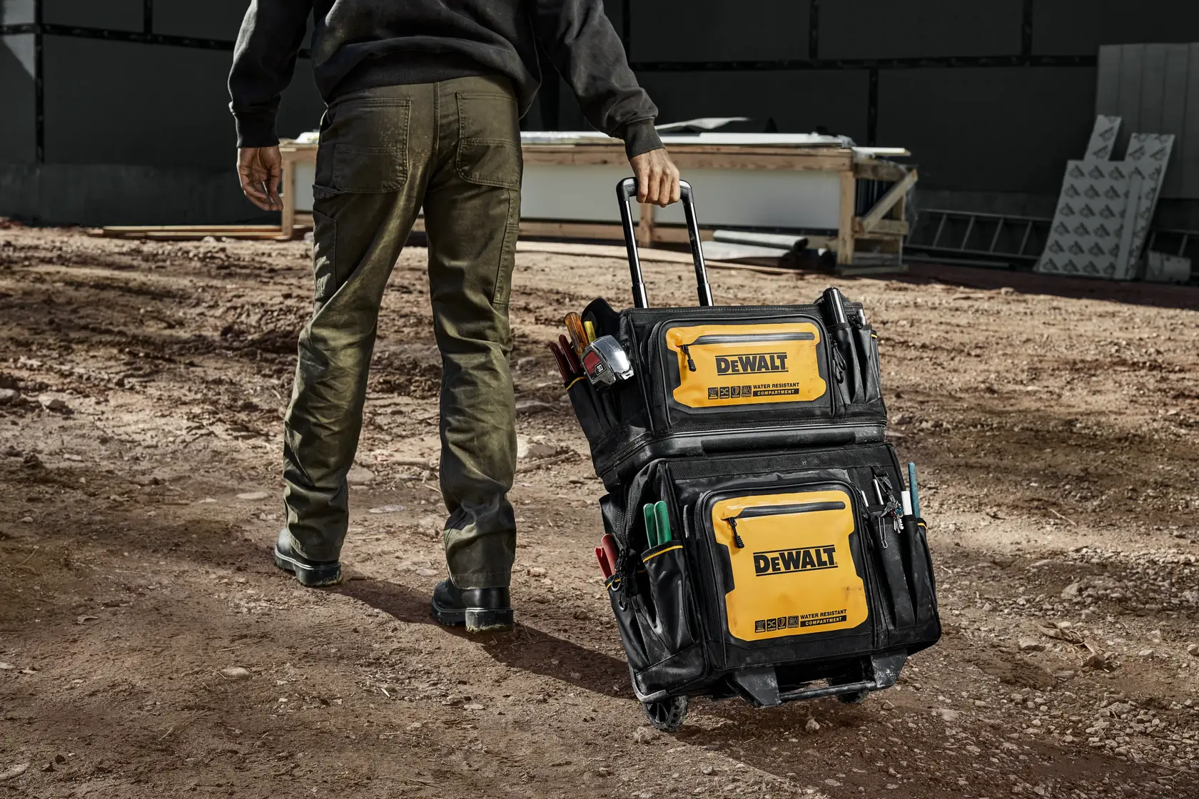 Uma pessoa está sobre uma superfície de cascalho e abre uma bolsa de ferramentas DEWALT, mostrando a grande abertura e compartimentos organizados com várias ferramentas, incluindo uma serra recíproca sem fio, alicates e marcadores. A bolsa destaca o fácil acesso às ferramentas.