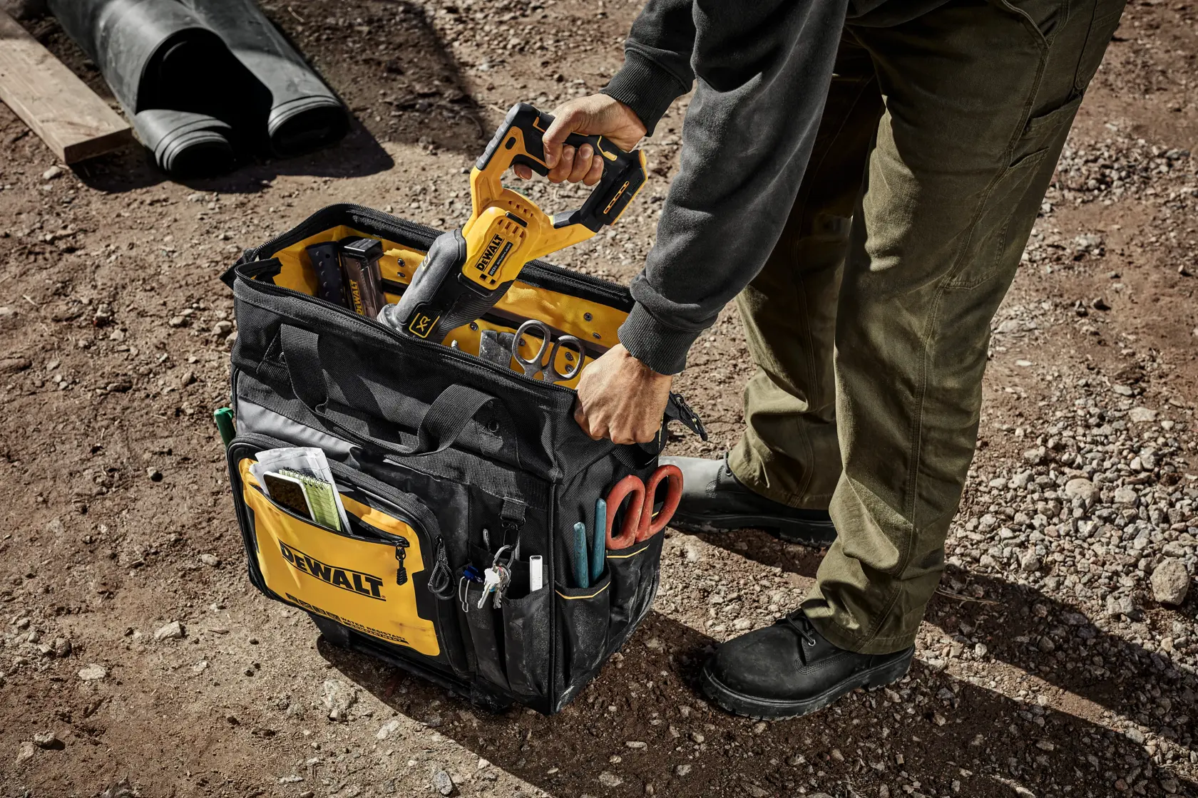 Uma pessoa está caminhando ao ar livre em um canteiro de obras, puxando uma bolsa de ferramentas com rodas da marca DEWALT cheia de ferramentas. Ao fundo, há um prédio parcialmente construído.