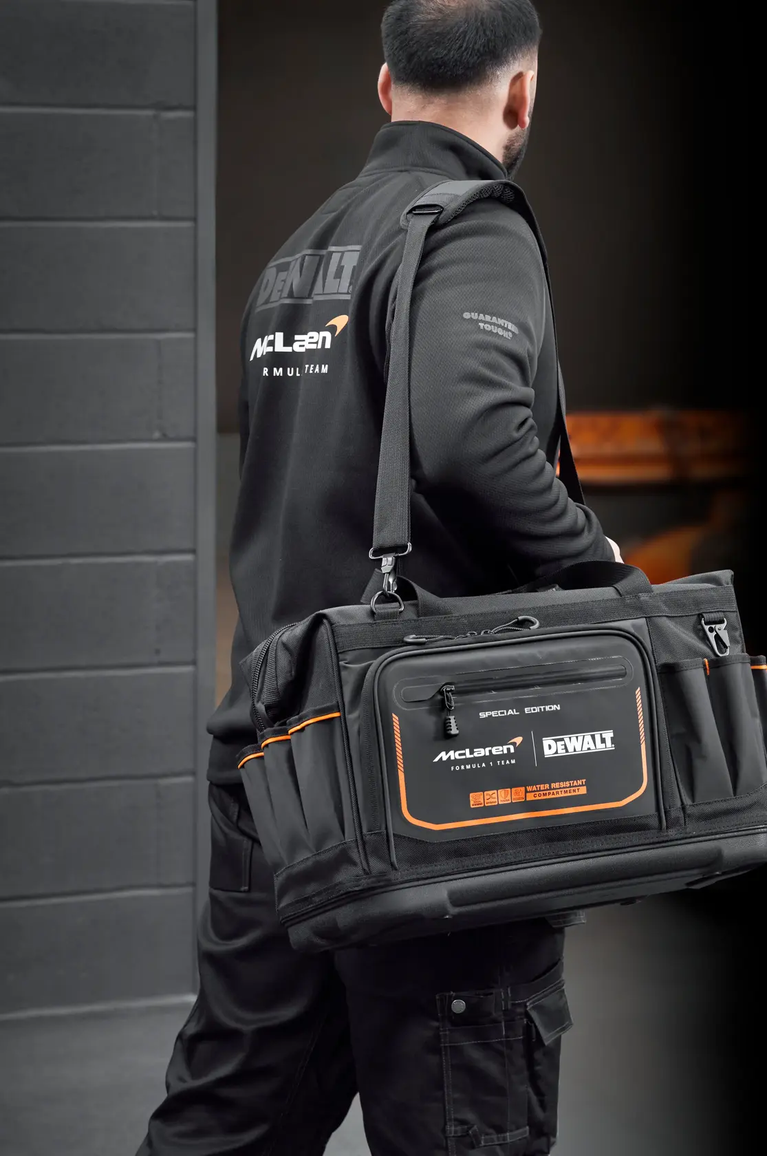 Pessoa carrega uma bolsa preta DEWALT McLaren com detalhes laranja em ambiente industrial.
