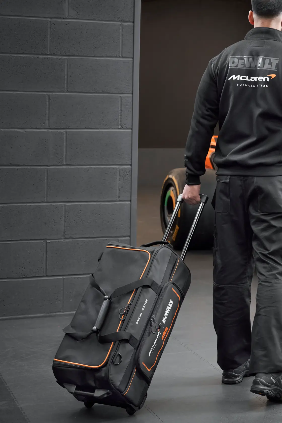 Pessoa puxa uma bolsa de ferramentas DEWALT McLaren com rodas em garagem com carro de corrida ao fundo.